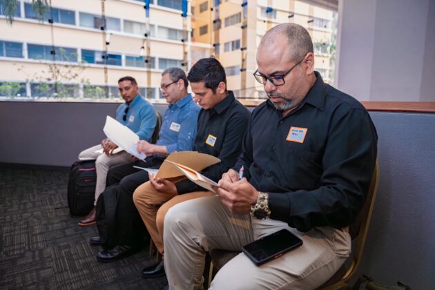 Four men sit on a bench and fill out a job application.