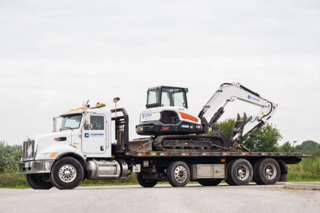 Large renter truck stands in a parking lot. It is carrying a construction machine for digging.